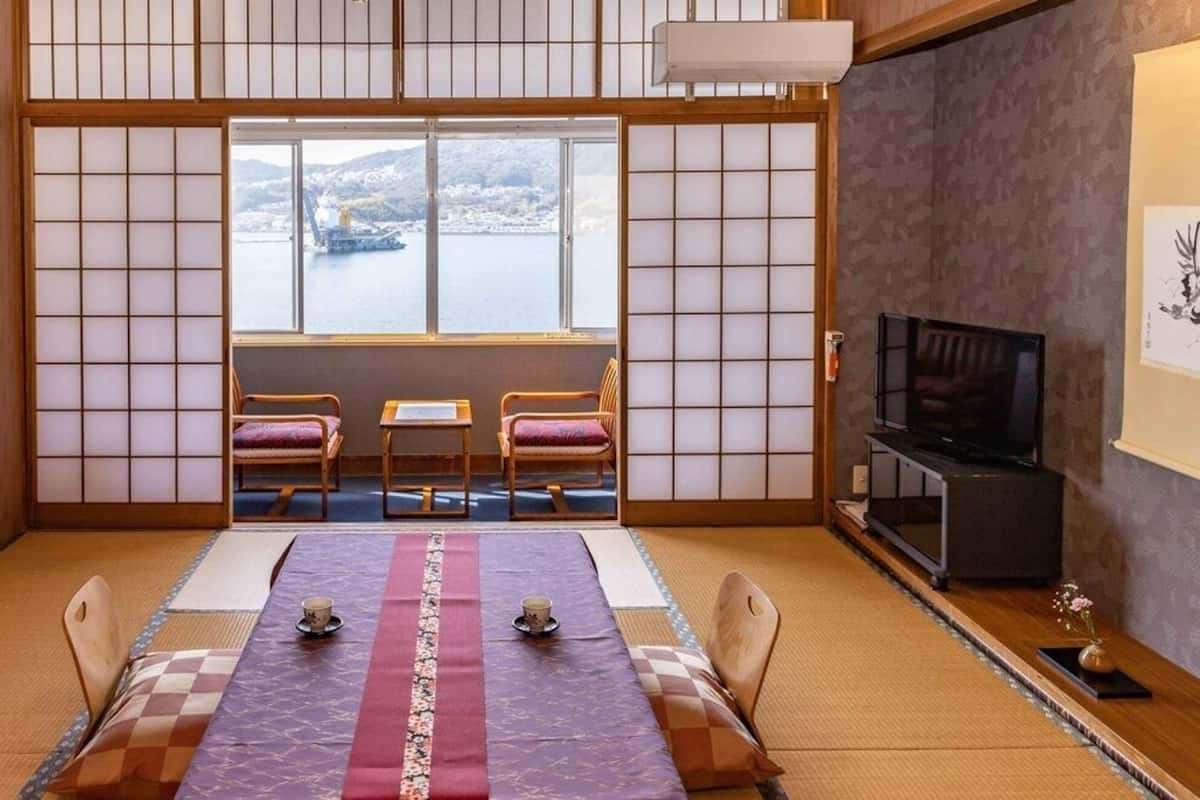 Ein traditioneller japanischer Raum mit Tatami-Böden, einem niedrigen Tisch mit Kissen, einem Fernseher und Blick auf ein Gewässer.