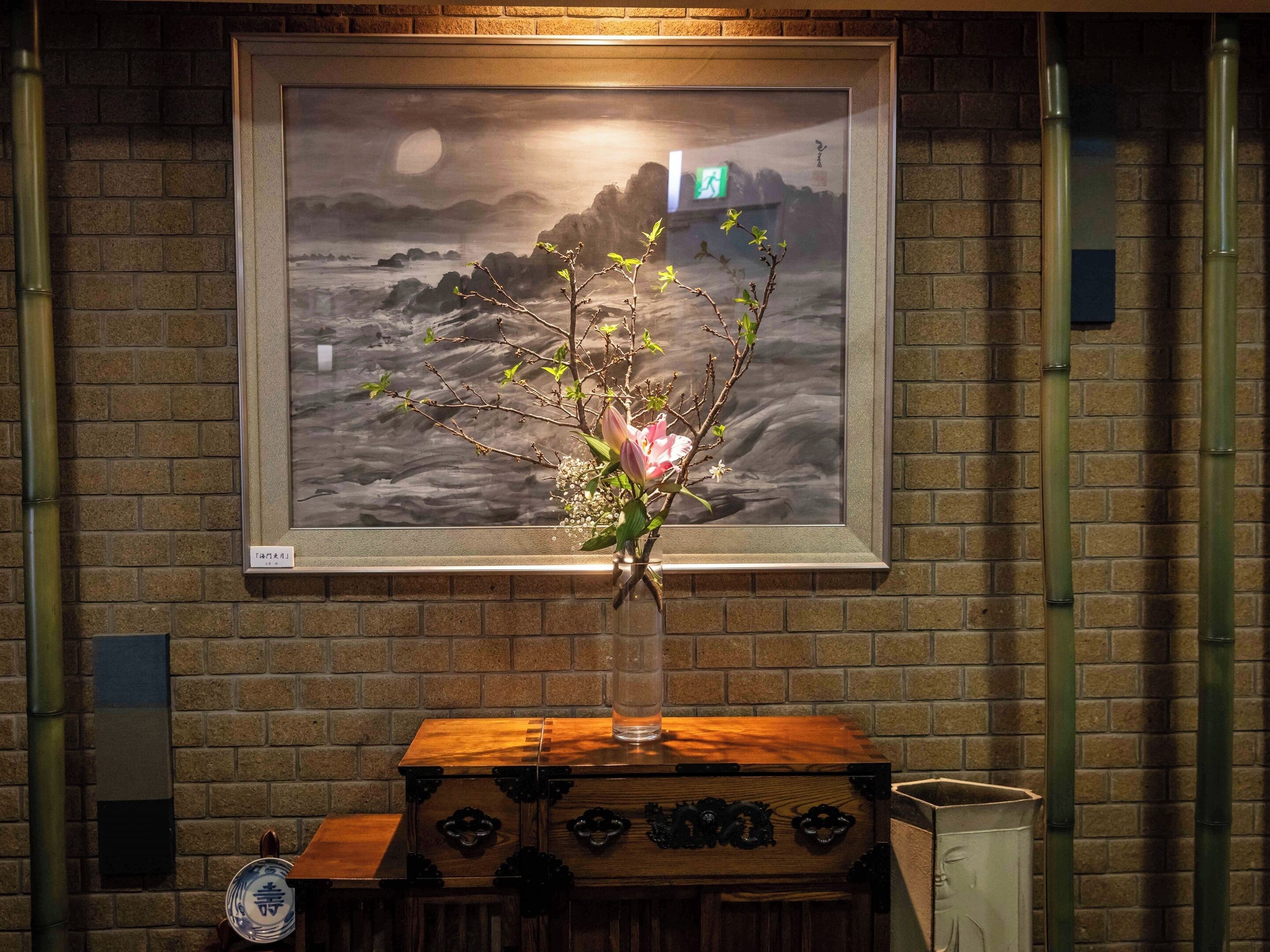 Una obra de arte enmarco que muestra un paisaje con luna y costa rocosa, un jarrón con flores y un cofre de madera con cajones.