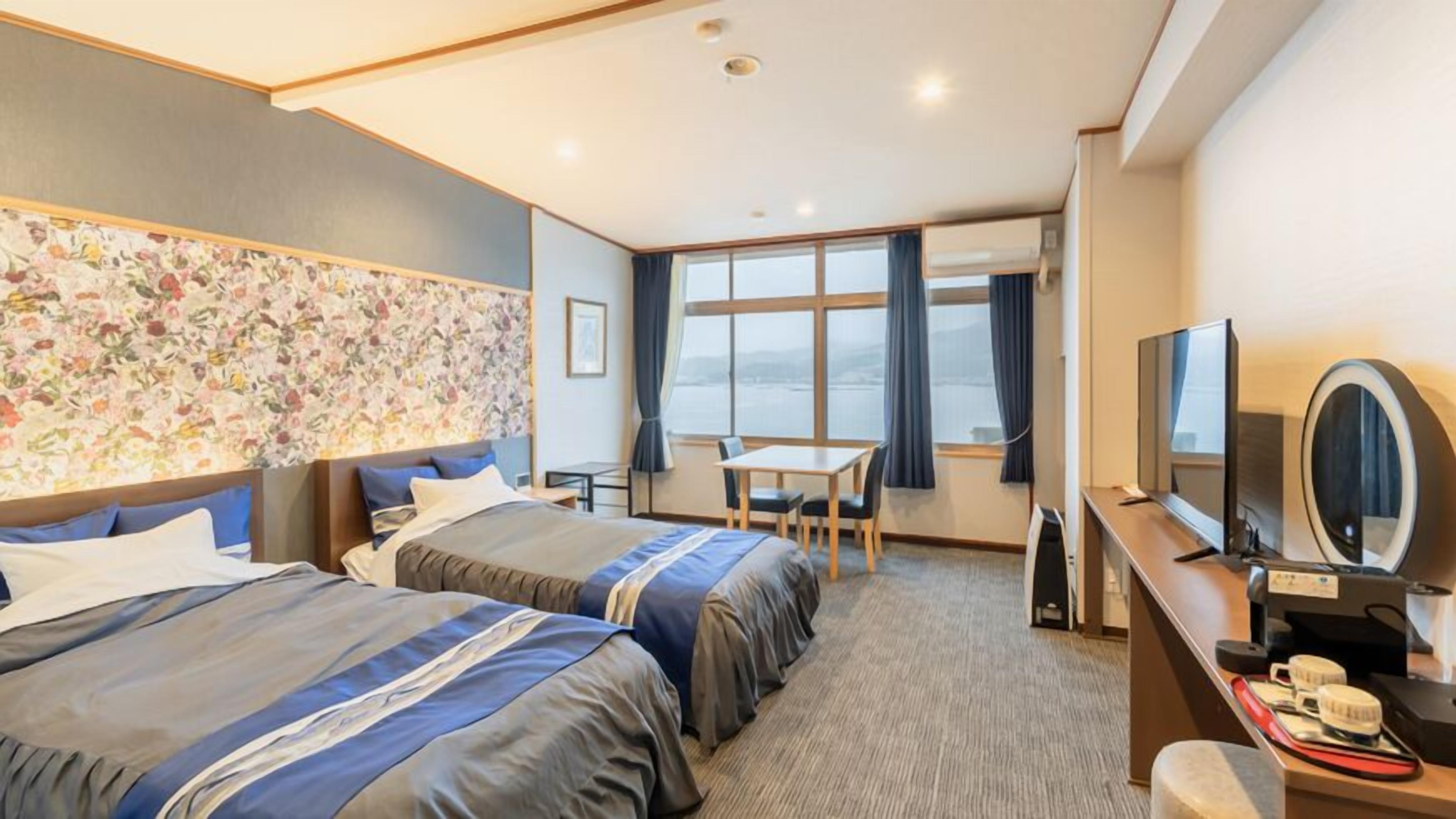 Habitación de hotel con dos camas, un escritorio, un televisor y un ventanal grande.