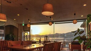 A restaurant with a view of a sunset over water, wooden furniture, and hanging pendant lights. - Shiozaki Onsen Awajishima kaijyo Hotel (Minamiawaji)