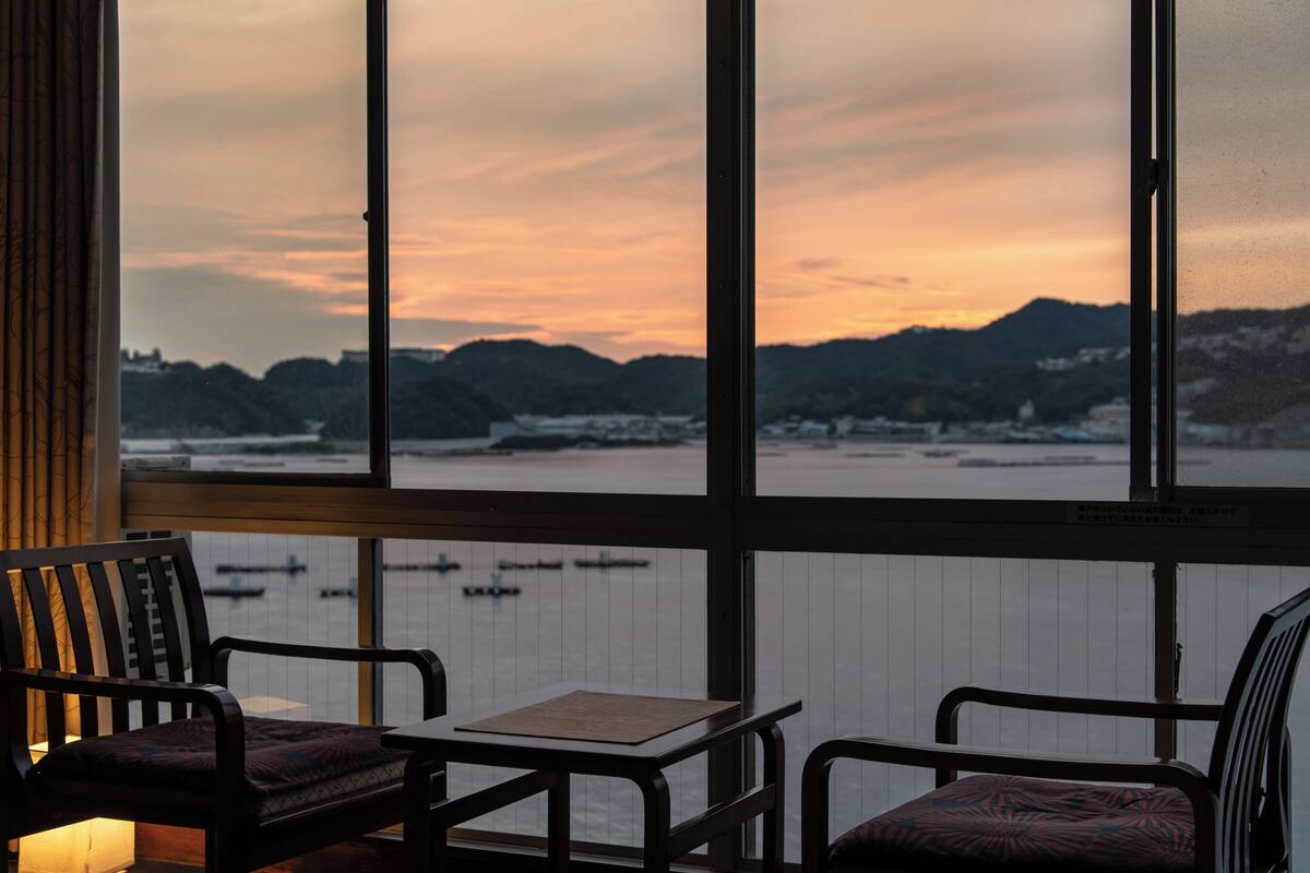 Ein Zimmer mit Blick auf einen Sonnenuntergang über einem Gewässer, ausgestattet mit zwei Stühlen und einem Tisch.