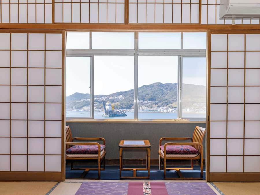 A traditional Japanese room with tatami flooring, a shoji screen, and a view of a harbor with mountains in the background.
