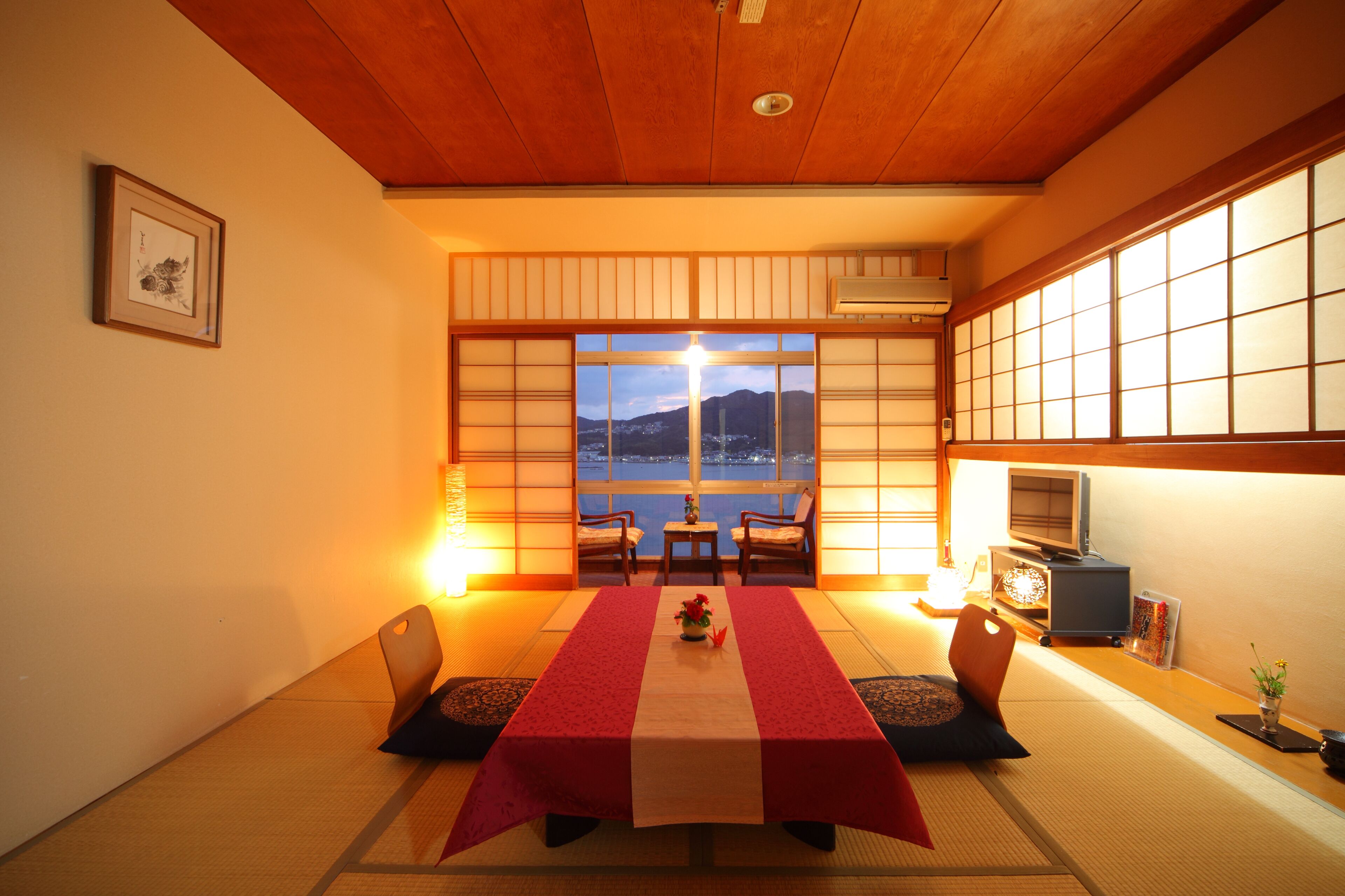 Une pièce de style japonais traditionnel, avec un sol recouvert de tatamis, une table basse recouverte d’une nappe rouge et blanche, et une vue sur un plan d’eau et des montagnes, visible à travers des paravents en papier.