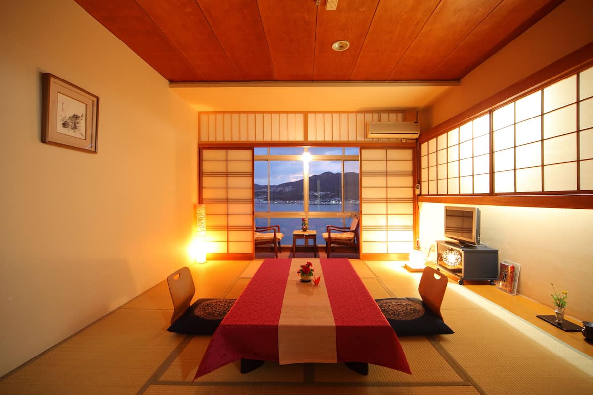 Une pièce de style japonais traditionnel, avec un sol recouvert de tatamis, une table basse recouverte d’une nappe rouge et blanche, et une vue sur un plan d’eau et des montagnes, visible à travers des paravents en papier.