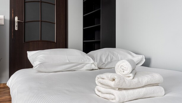 A neatly made bed with white linens and pillows, a dark wooden door with a glass panel, and a black shelving unit against the wall.