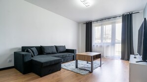 Un salon moderne avec un canapé d’angle, une petite table en bois et une grande fenêtre ornée de rideaux légers.