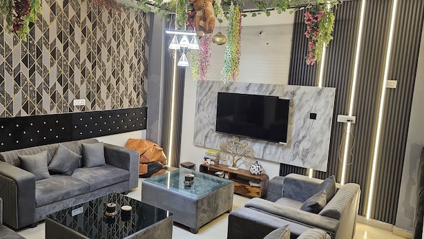 A modern living room with a grey sofa, a glass coffee table, and a flat-screen TV mounted on a marble wall. - Bunk house villa (Noida)