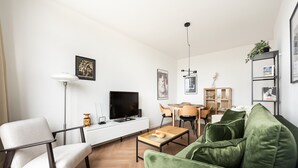 Een moderne woonkamer met een groene bank, een witte fauteuil, een televisie, een eettafel met stoelen en een boekenkast.