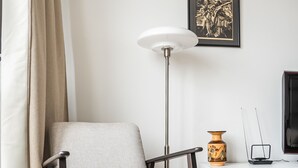 Een moderne woonkamer met een lichtkleurige fauteuil, een vloerlamp, een witte bijzettafel met een vaas en een klein sculptuur, en een kunstwerk in een lijst aan de muur.