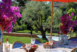 An outdoor dining area with a table set for two, surrounded by lush greenery and a pool in the background.