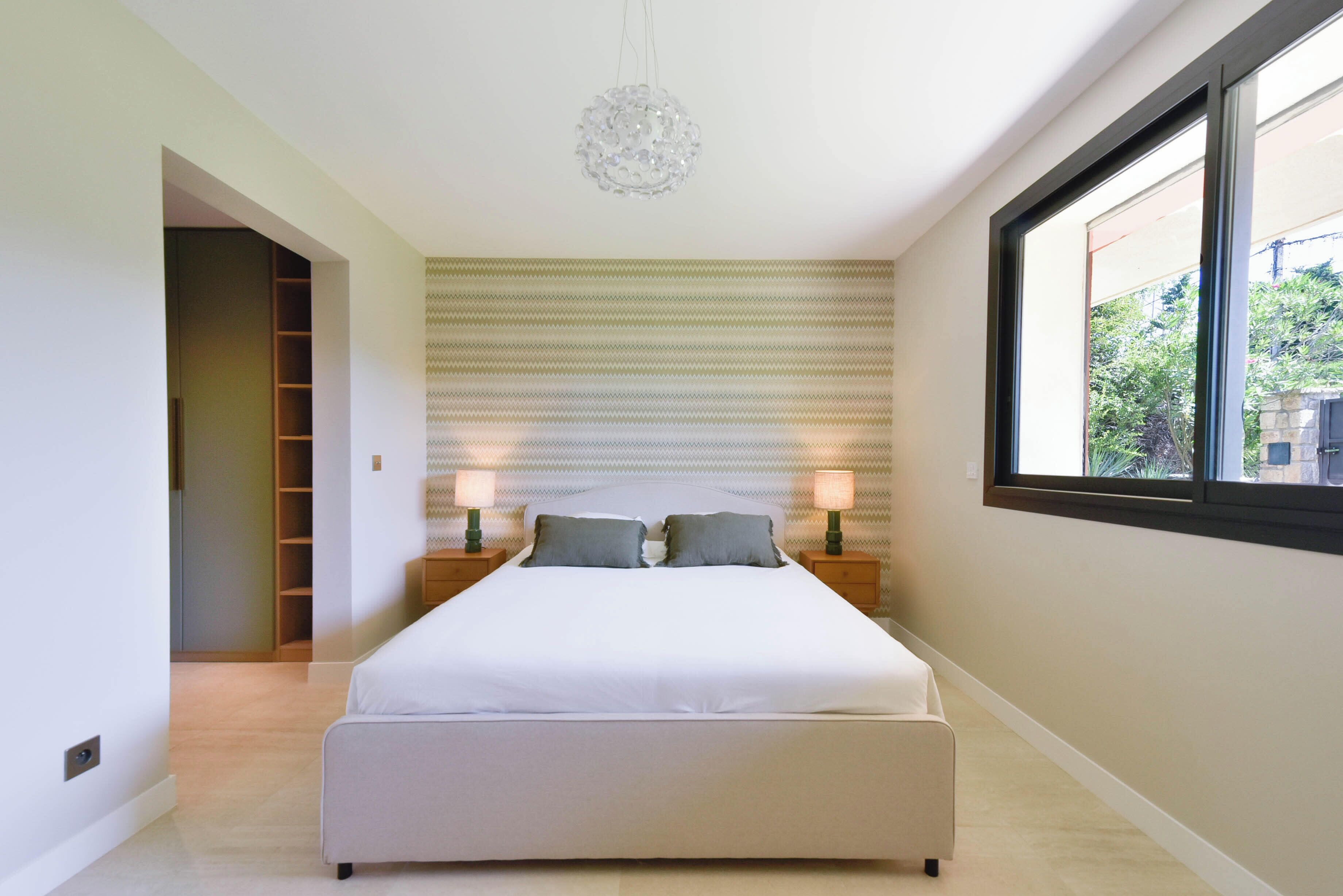 A bedroom with a large bed, two bedside tables with lamps, a patterned wall, and a window with a view.