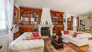 Un vaste salon agrémenté de deux canapés en cuir blanc, d’une cheminée et de grandes étagères en bois remplies de livres et d’objets décoratifs.