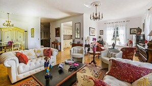 Un salon spacieux comprenant un canapé, des fauteuils, une table basse et un lustre. On y trouve également un coin repas avec une table et des chaises, ainsi qu’une cuisine à l’arrière-plan.