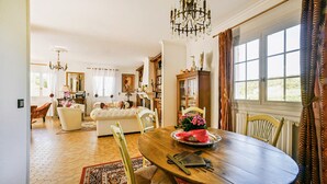 Un coin repas avec une table ronde en bois, des chaises et un lustre. Une grande fenêtre, ornée de rideaux, offre une vue sur la verdure environnante.