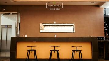A reception desk with three stools, a window with curtains, and a sign above stating "Plaza Acequia Suites & Rooms".