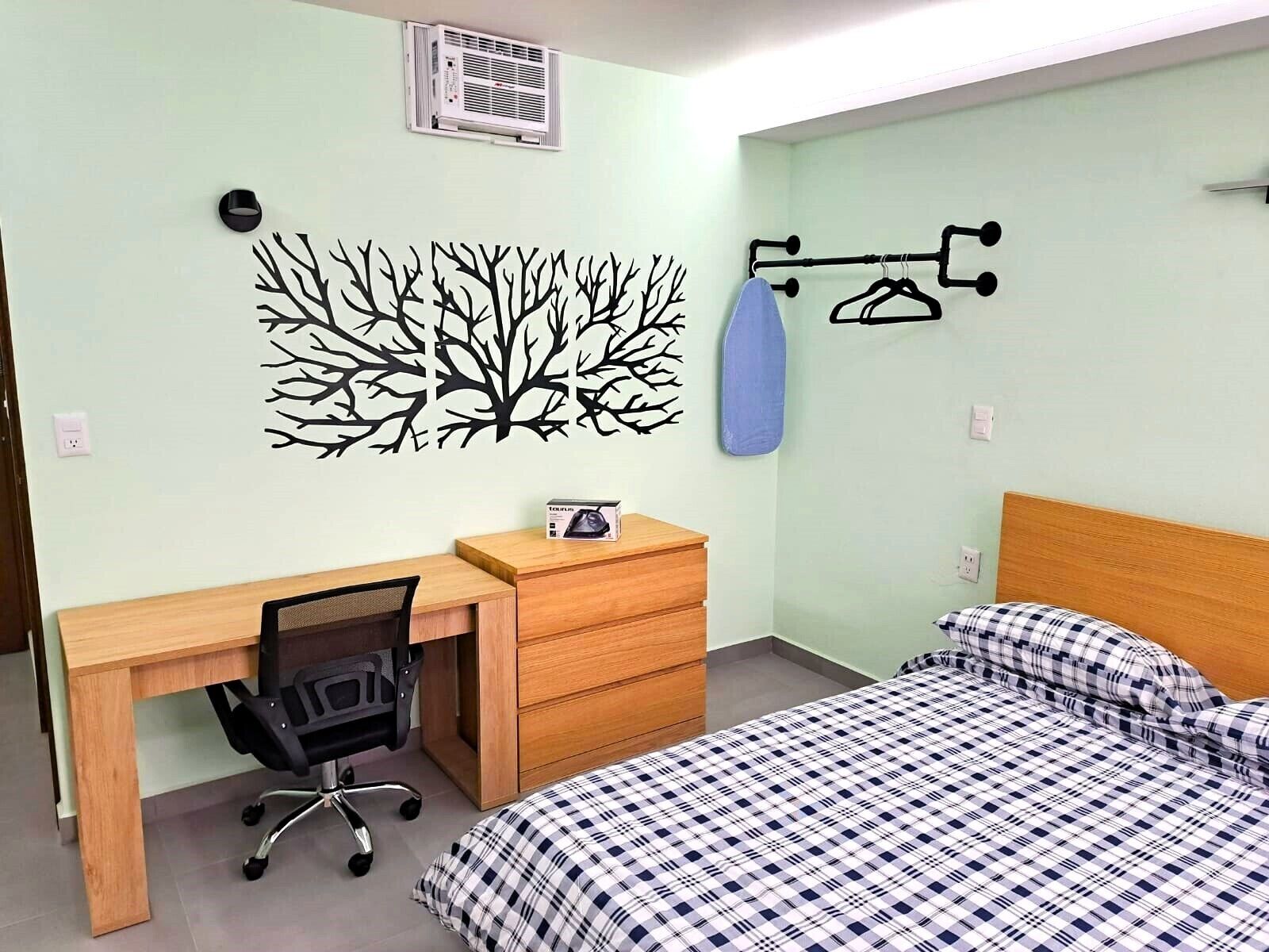 Une chambre avec un bureau en bois, une chaise, un lit avec une couette à carreaux, un climatiseur fixé au mur et une décoration murale représentant des arbres.