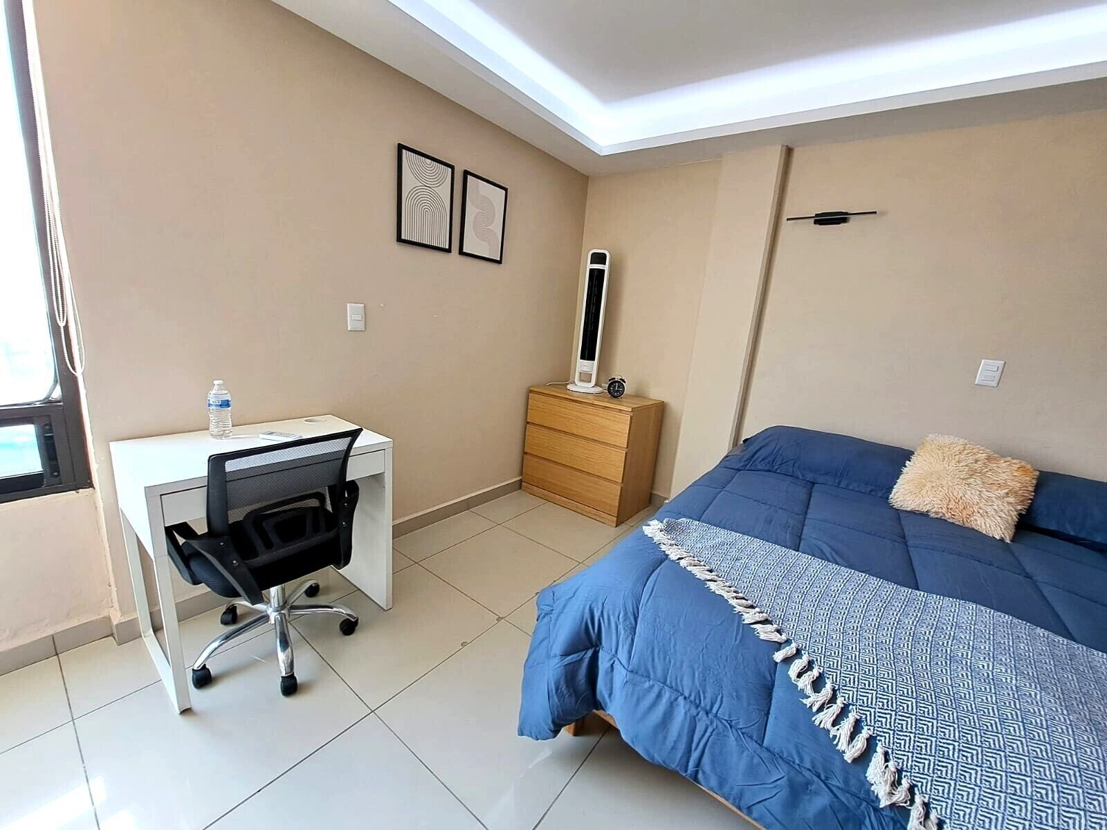 A bedroom with a bed, a desk, a chair, a dresser, and a bedside table with a lamp.