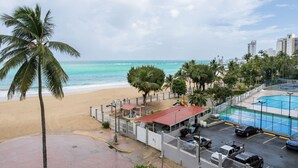 Vista de una playa con zona de piscina, palmeras y un estacionamiento cercado.