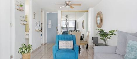Una sala de estar con un sillón azul, un sofá gris, un ventilador de techo y un comedor al fondo.