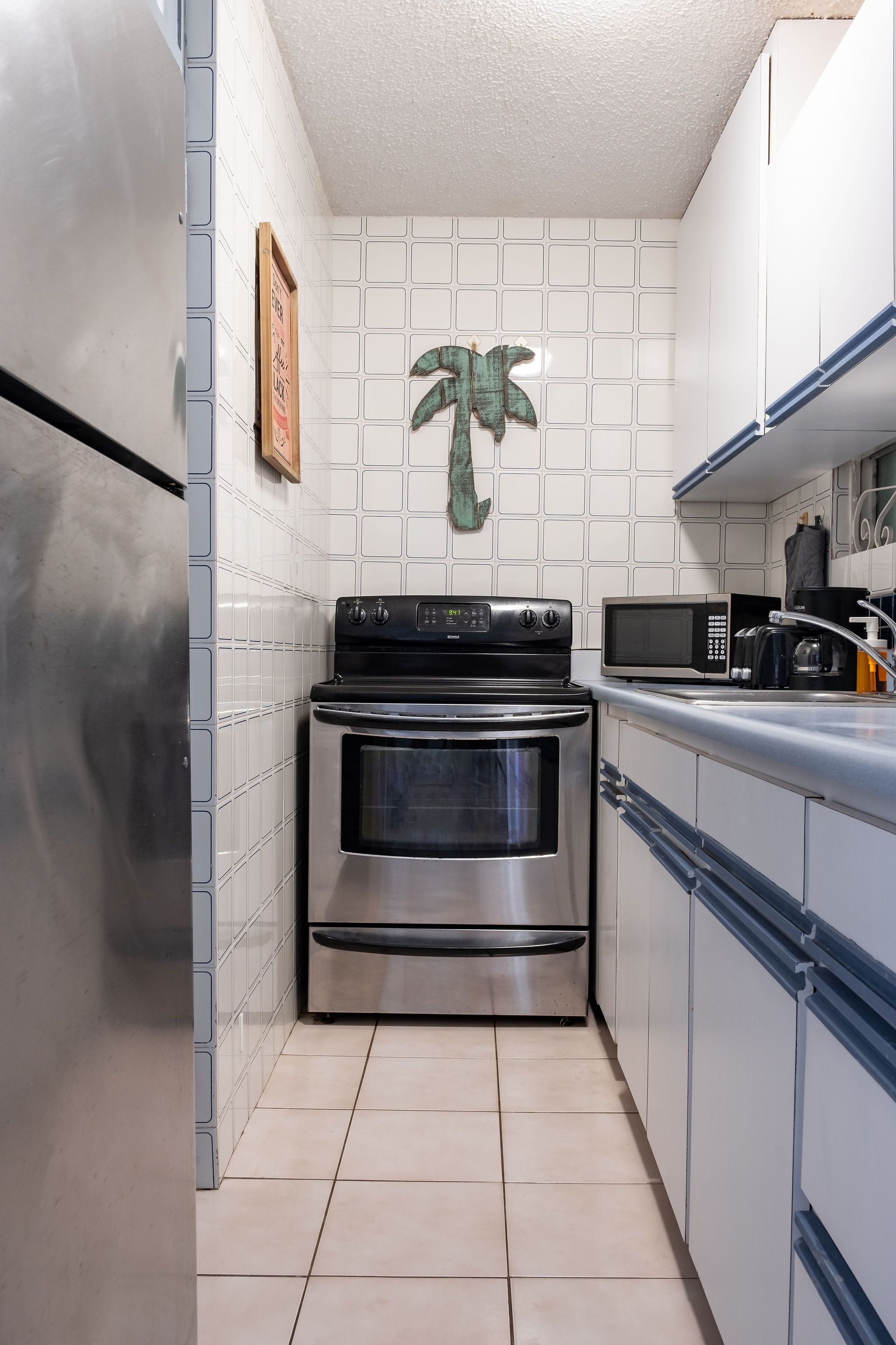 A compact kitchen with stainless steel appliances, white tiled walls, and a decorative palm tree on the wall.