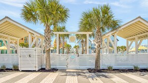 A gated entrance with palm trees, a covered walkway, and buildings in the background. - Resort Style Pools, Golf Cart Boardwalk to the Beach, Pet Friendly (Port Aransas)