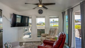 Un salón con ventilador de techo, televisor de pantalla plana, un sillón de cuero rojo, un sofá color beige y ventanas grandes que ofrecen vistas a la playa y montañas.