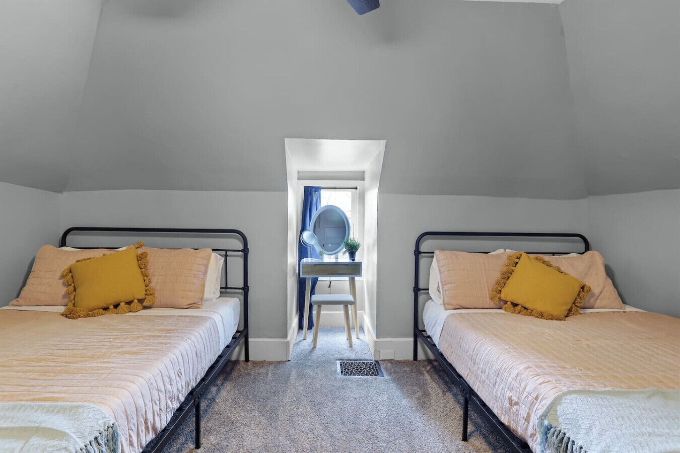 A bedroom with two beds, each with a headboard, a small desk with a mirror, and a window with curtains.