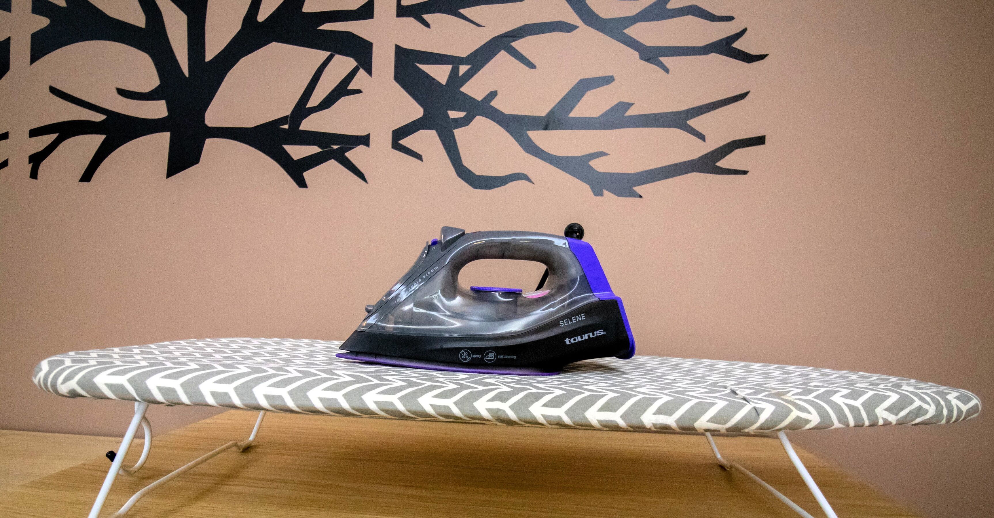 An ironing board with an iron on it, a patterned wall, and a wooden table.