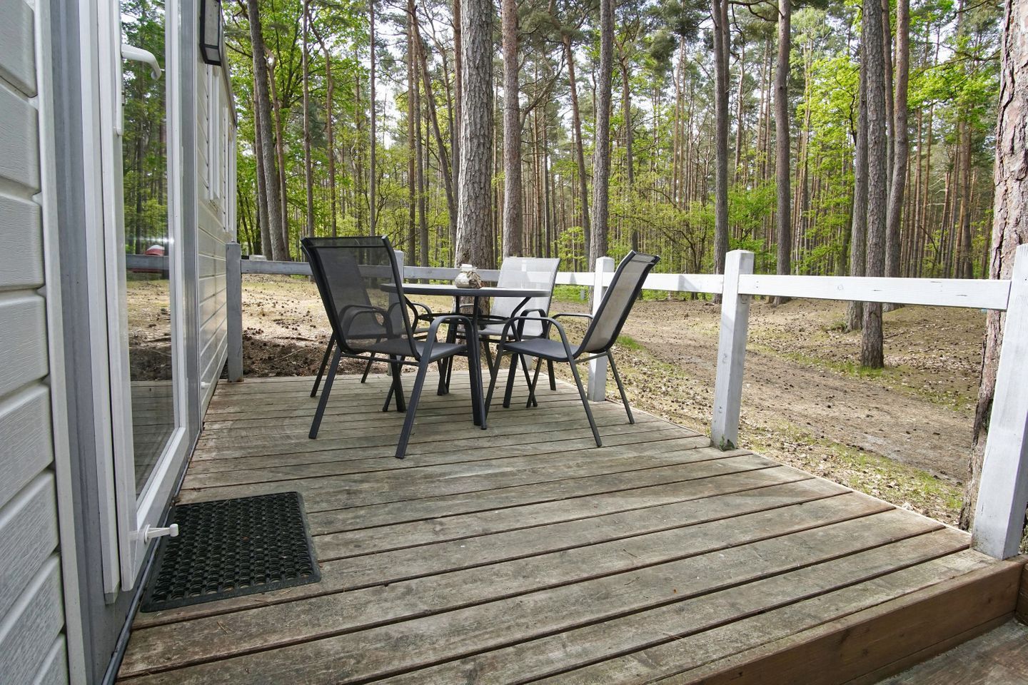 Eine Holzterrasse mit Tisch und Stühlen, umgeben von einem weißen Zaun und einem bewaldeten Gebiet.