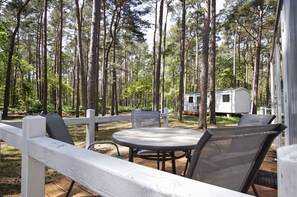 A covered outdoor seating area with a table and chairs, surrounded by a white fence, in a forested setting with a mobile home in the background. - Kleines Aber Feines Ferienhäuschen in Strandnähe - Tiny Chalet Nr. 818 - Tiny Chalets (Binz)