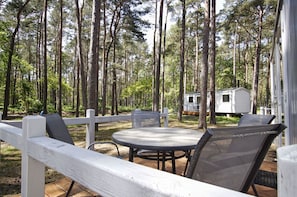 Et overdækket udendørs opholdsområde med et bord og stole, omgivet af et hvidt hegn, i en skovagtig omgivelser med en campingvogn i baggrunden.