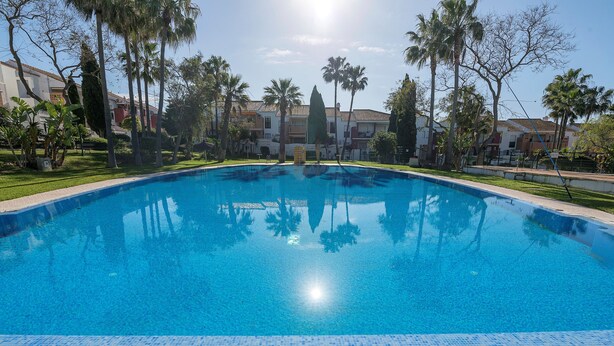 A clear blue swimming pool surrounded by palm trees and a building with a tiled roof. - OASIS PLAYERO - Nice flat with shared pool and Free Wifi (Chiclana de la Frontera)