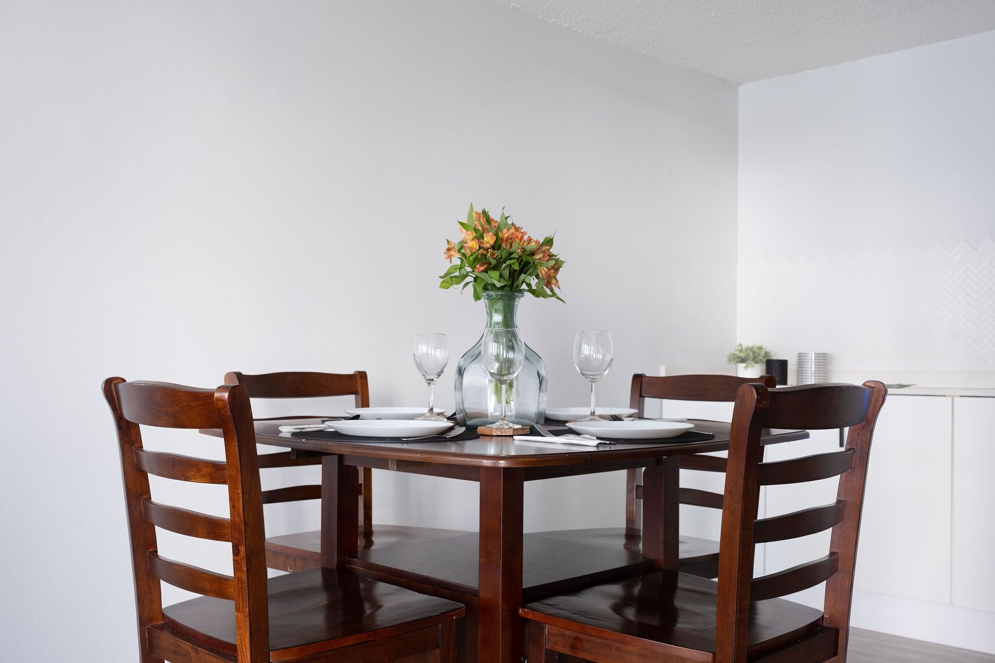 A dining table with four wooden chairs, a vase with flowers, and wine glasses set for four.