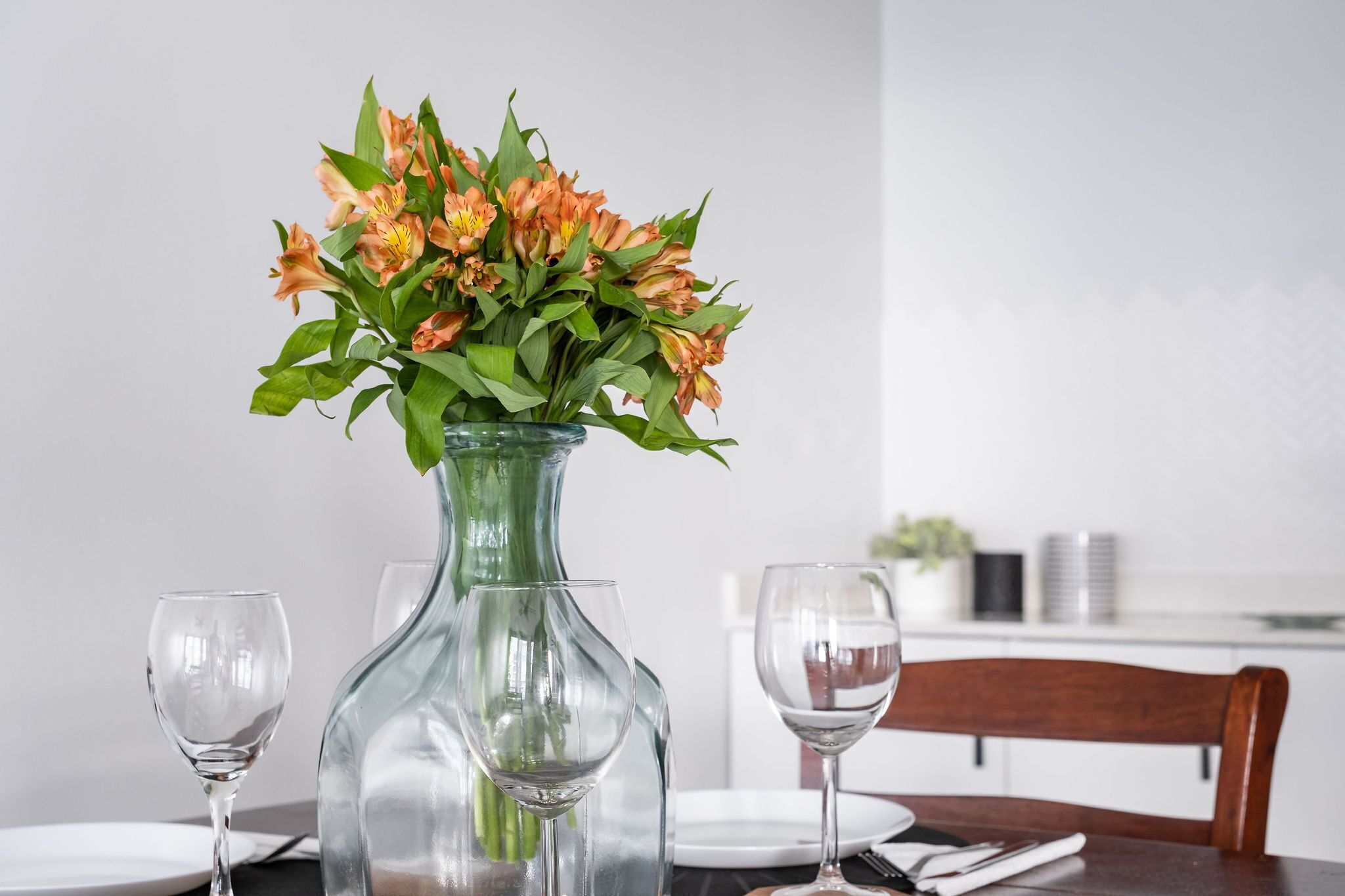 Une table à manger sur laquelle se trouve un vase en verre transparent contenant des fleurs oranges, deux verres à vin vides et une chaise en bois.