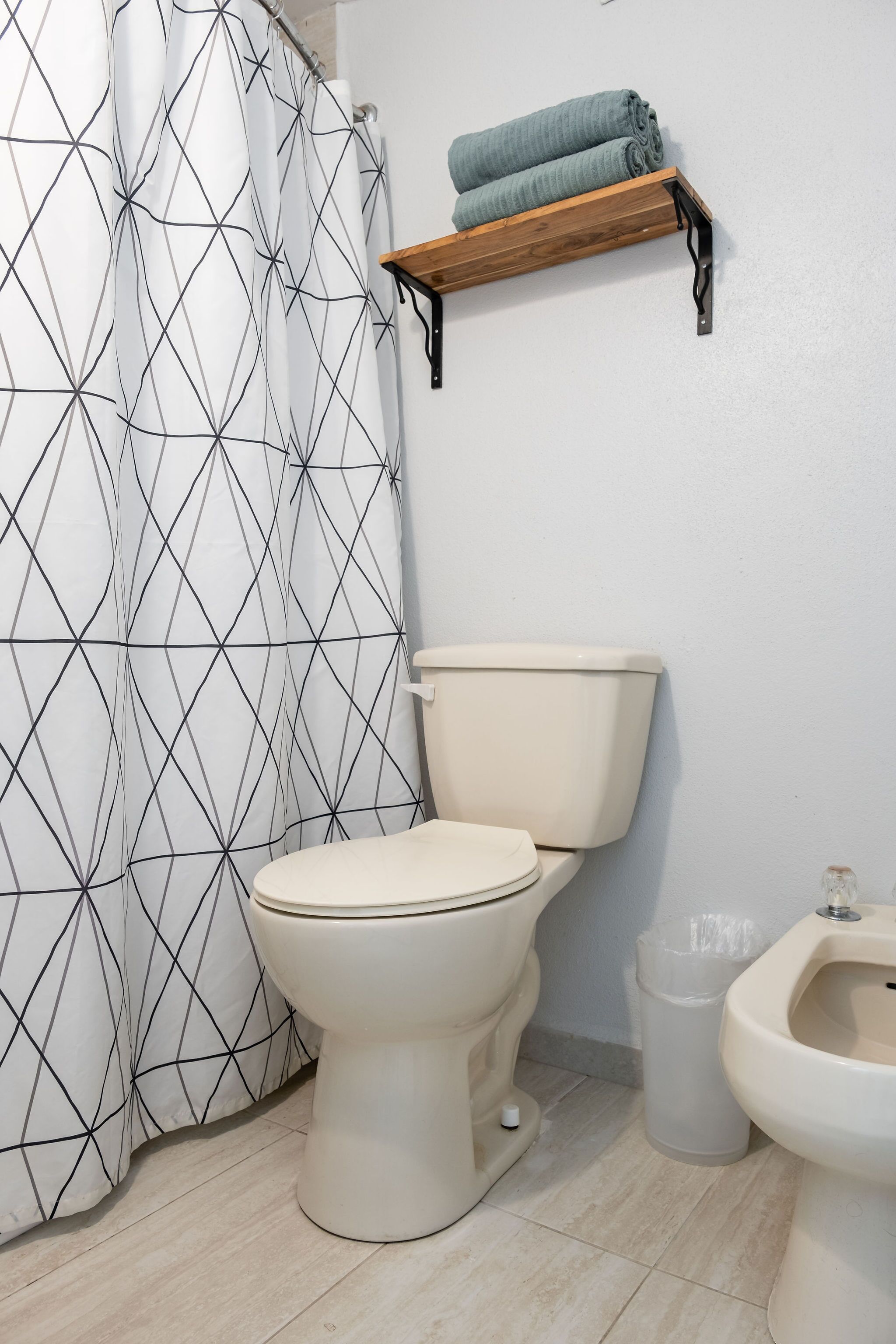 Un baño con inodoro, bidé, cortina de ducha con diseño geométrico y estante de madera con toallas.