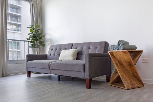 A grey sofa with a cushion, a wooden side table, and a potted plant in a room with a view of a building through a window.