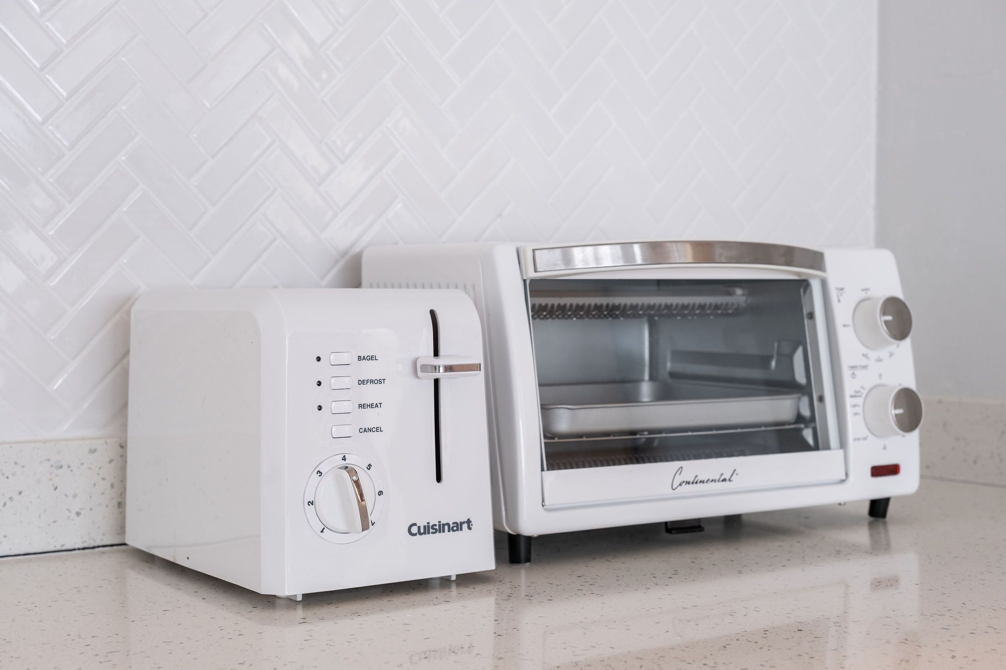 A white Cuisinart toaster oven with a control panel and a glass door, placed on a tiled floor against a white herringbone-patterned wall.