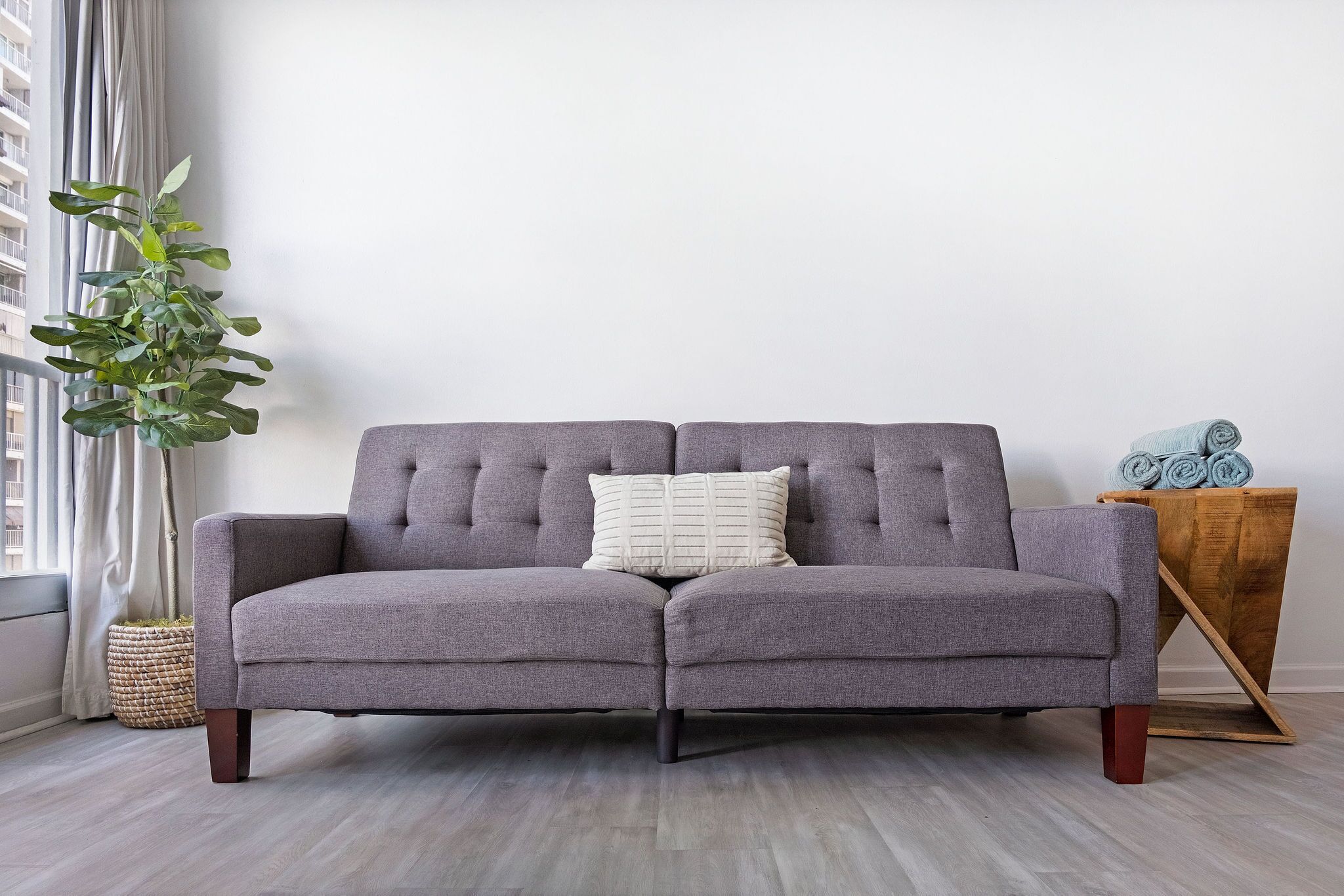 A grey sofa with a tufted backrest and a single white pillow, positioned against a plain wall with a window on the left.