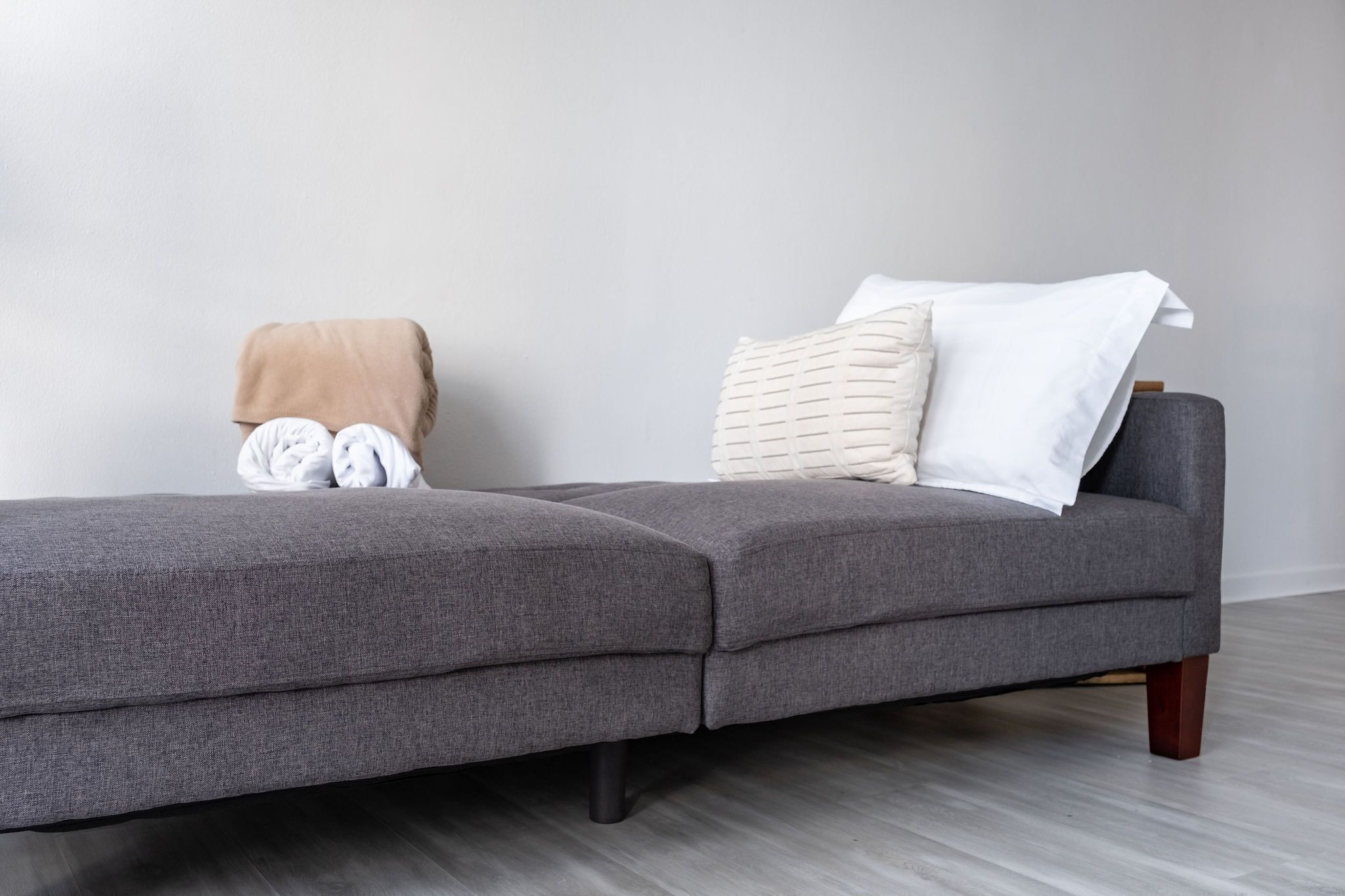 A grey sofa with a beige blanket and two pillows, a wooden floor, and a plain wall.