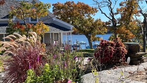 Una casa con porche cubierto, un jardín con diversas plantas y flores, y vista al agua con barcos al fondo.