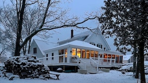 Una casa grande y blanca con un porche envolvente y una chimenea, rodeada de árboles cubiertos de nieve.