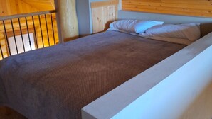 Een kamer met een enkel bed, houten plafond en muren, een wit hoofdbord en een grijze dekbedovertrek.