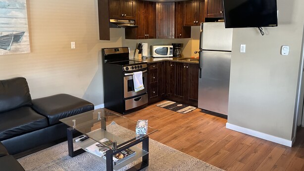 Een compacte leefruimte met een kitchenette, een flatscreen-tv aan de muur, een glazen salontafel en een donkergrijze bank.