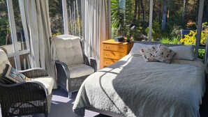 A bedroom with a large bed, a nightstand, a dresser, and two chairs. There is a window with curtains, and a view of greenery outside.