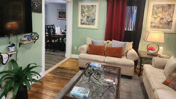 A living room with a sofa, coffee table, and a television. There is a dining area visible through an open doorway.