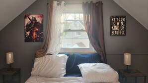 A bedroom with a sloped ceiling, a bed with white and blue bedding, a window with curtains, a wall-mounted sign, and a lamp on a bedside table.