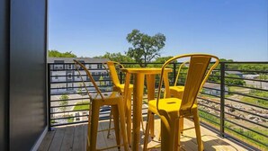 A balcony with yellow chairs and a table, overlooking a parking area and a tree.