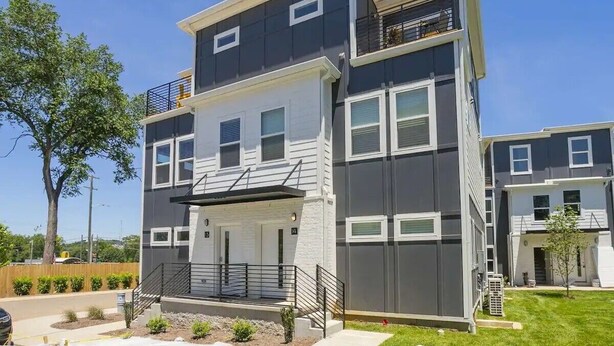 A modern multi-story residential building with a balcony on the upper floor, a covered entryway, and a visible front door.