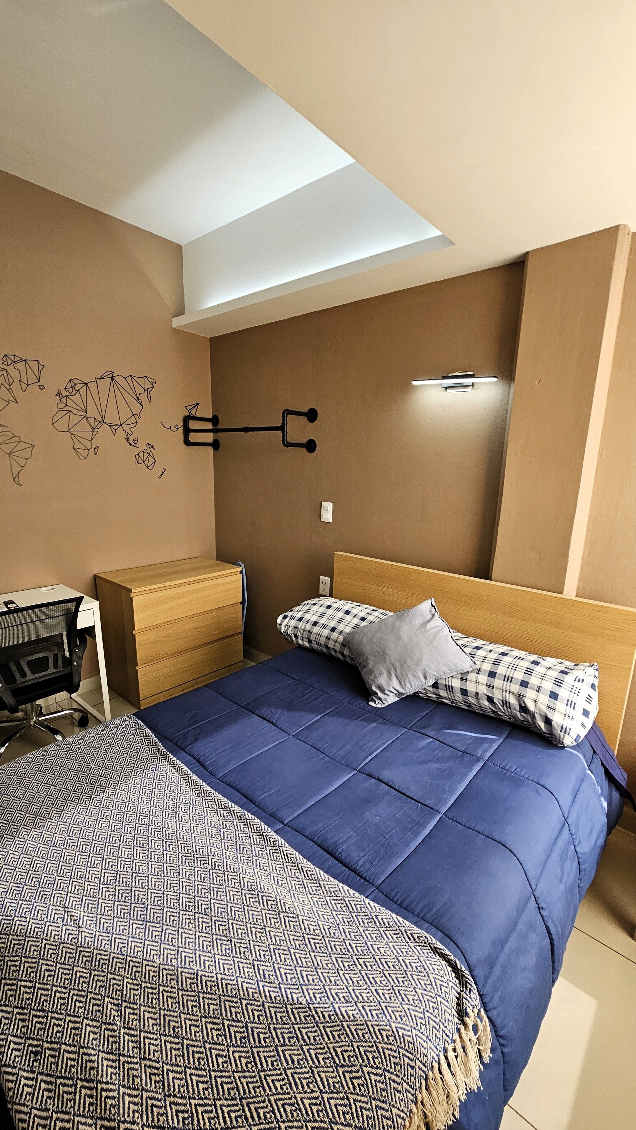 A bedroom with a bed, a desk, and a chair. There is a map on the wall and a light fixture above the bed.