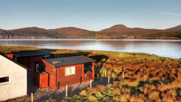 A cabin with a small attached structure, a fenced yard, and a view of a lake and hills in the background. - Harlosh Log Cabins, Cabin No 1 (Dunvegan)